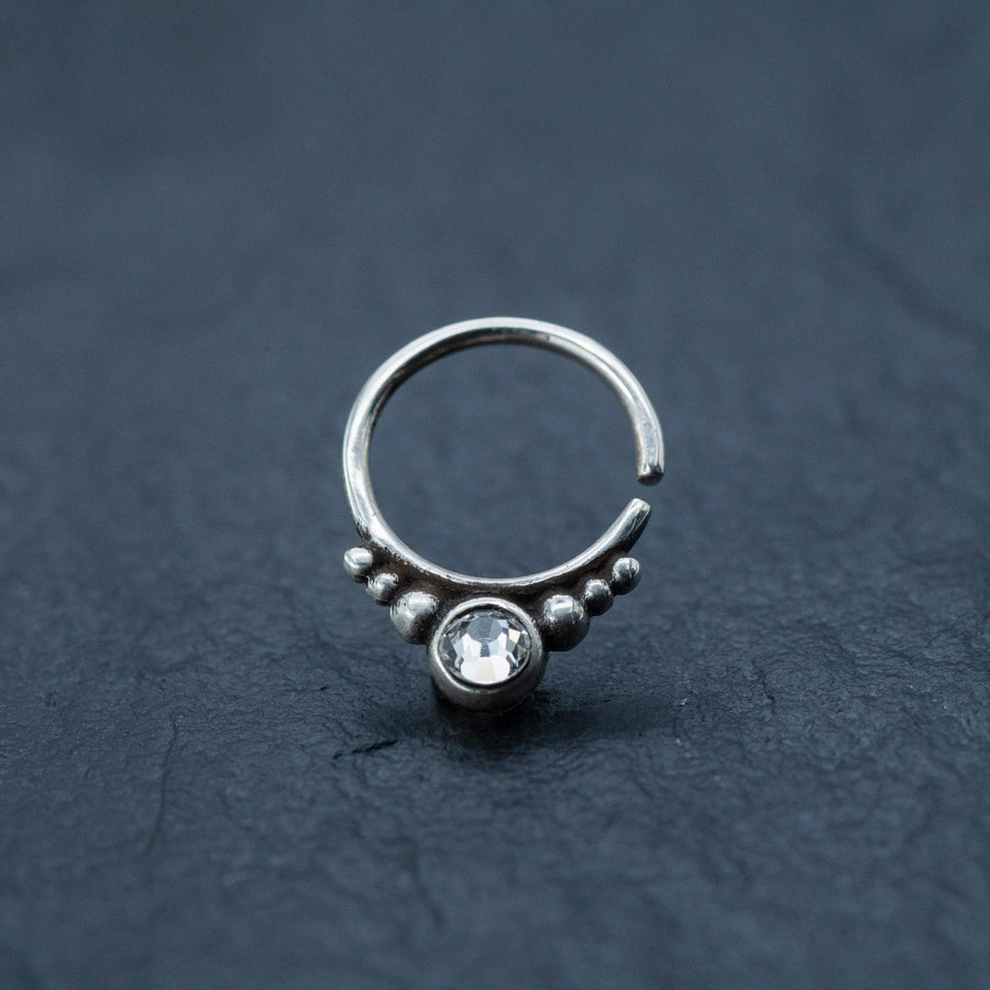 Close-up of the Zircon Silver Nose Septum Ring Piercing by Kaya Movement, showcasing its unique circular design. This sterling silver piece features a prominent central zircon gem, surrounded by small spherical accents on a dark, textured surface. The cute 8mm teardrop shape adds a touch of goth-inspired elegance to this daith jewelry.
