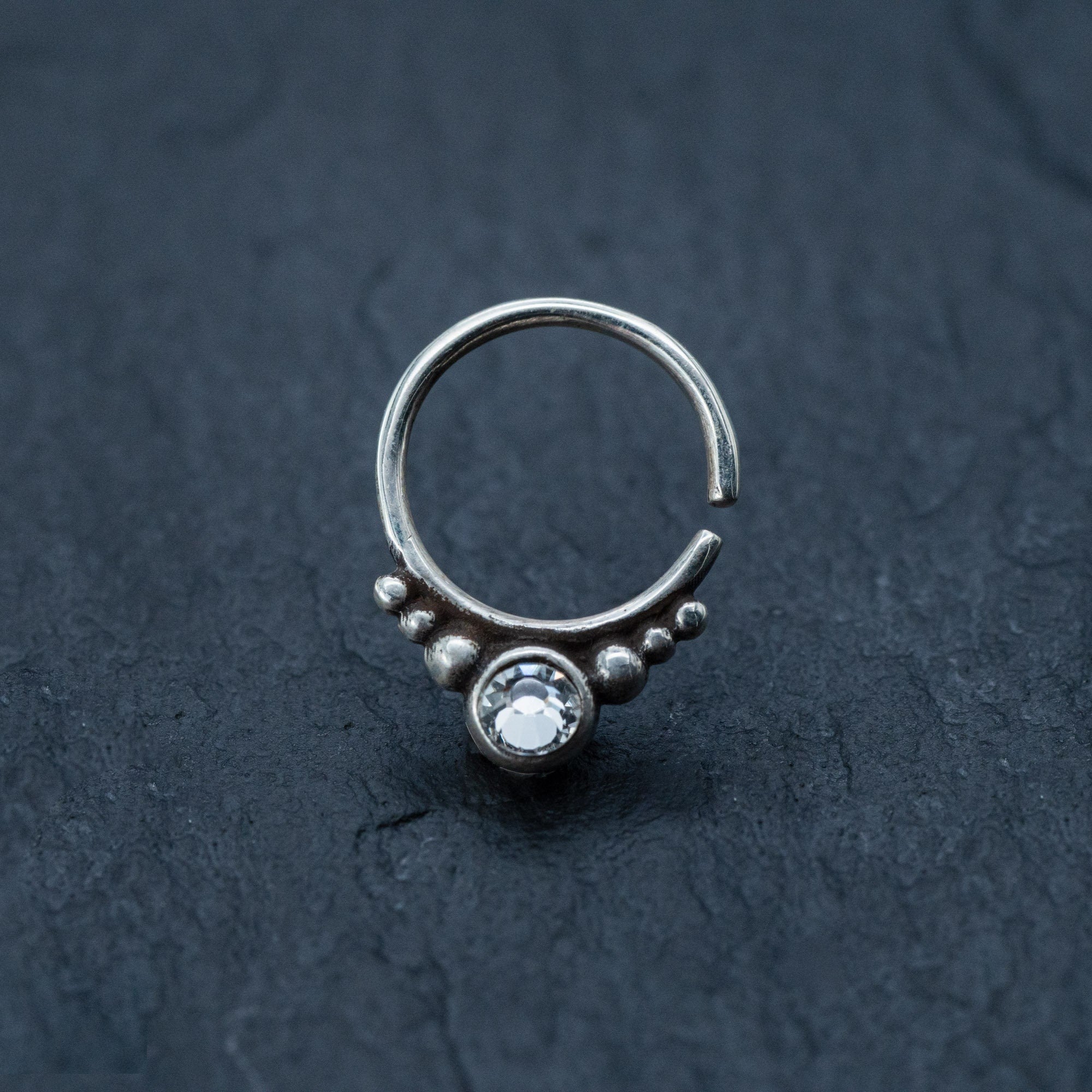 The Zircon Silver Nose Septum Ring Piercing by Kaya Movement, a small, teardrop-shaped septum nose ring with a decorative design, is shown on a dark textured surface. The ring features a clear, round gemstone at the bottom, flanked by a row of small silver beads, adding an intricate and elegant touch.