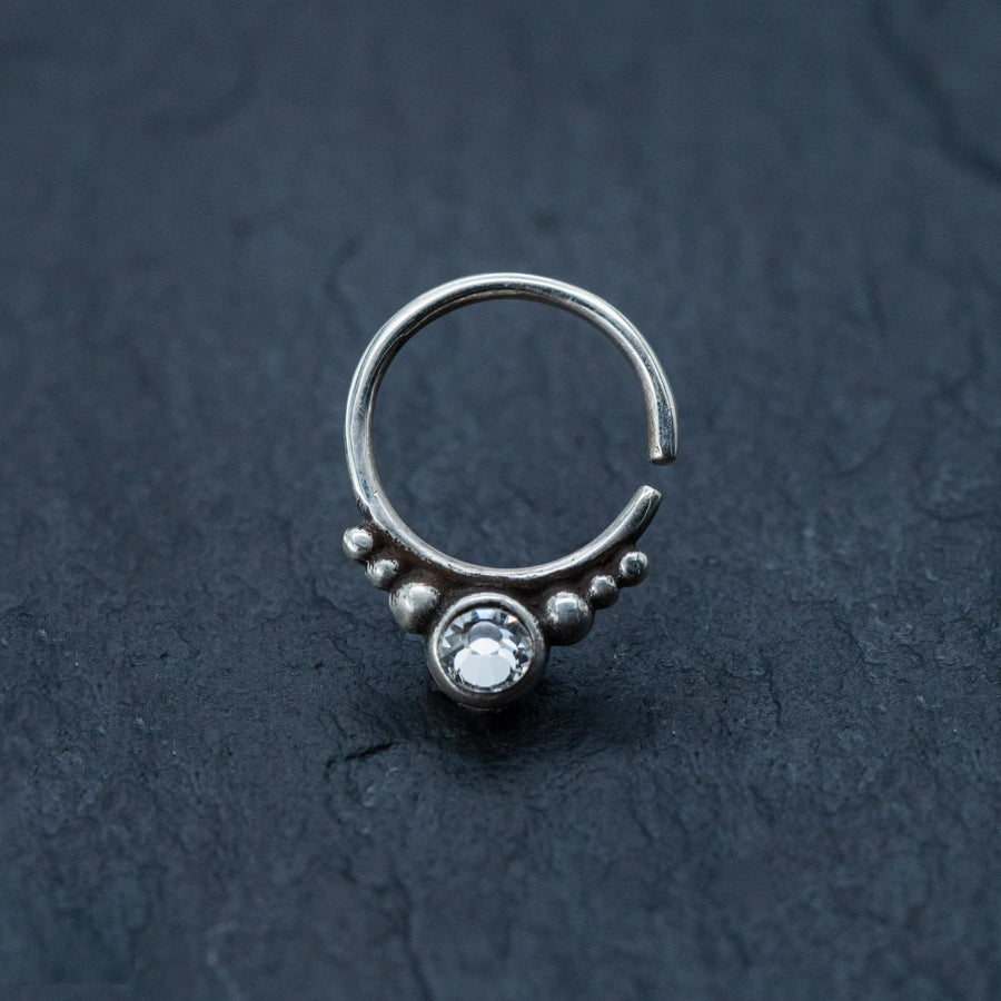 The Zircon Silver Nose Septum Ring Piercing by Kaya Movement, a small, teardrop-shaped septum nose ring with a decorative design, is shown on a dark textured surface. The ring features a clear, round gemstone at the bottom, flanked by a row of small silver beads, adding an intricate and elegant touch.
