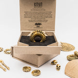 A sterling silver septum ring featuring turquoise stone and a bohemian alternative design is presented inside an open cardboard box with black padding. The box lid is inscribed with "Kaya Movement." Surrounding the box are various ornate gold jewelry pieces, including snakes and skulls, alongside elements of bohemian fashion.