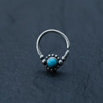 A close-up image of the Kaya Movement Silver Septum Ring with a small turquoise gemstone at the center, surrounded by beaded detailing. The background is a dark, textured surface.