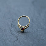 The Kaya Movement Garnet Teardrop septum, an 8mm seamless gold nose ring, features a delicate circular design. This elegant brass piece is embellished with a small red gemstone at the front and flanked by tiny bead-like gold accents. Perfect as a garnet nose piercing ring or for rook and daith piercings, it is showcased against a dark, textured surface background.