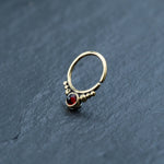 A delicate brass body jewelry piece from Kaya Movement, the Garnet Teardrop septum features a red gemstone centerpiece and decorative gold beads, elegantly sitting on a dark, textured surface. This seamless 8mm gold septum ring is perfect for rook or daith piercings.