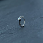 A Kaya Movement 925 silver nose ring with a small, round opal gemstone rests on a textured dark gray surface. The design features an 8mm septum and includes a band adorned with numerous small, decorative bumps leading up to the gemstone. Perfect for tragus, rook, or helix piercings, this elegant and minimalist silver nose hoop is ideal for lovers of opal jewelry.