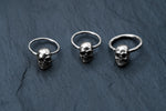 Three Kaya Movement Skull Ball Closure Rings, crafted from Stainless Steel 316L, are displayed on a dark, textured surface. The silver punk Goth hoop earrings with charm are aligned in a row, showcasing the intricate detailing on the skulls. The background contrasts with the metallic shine of the rings, highlighting their features beautifully.