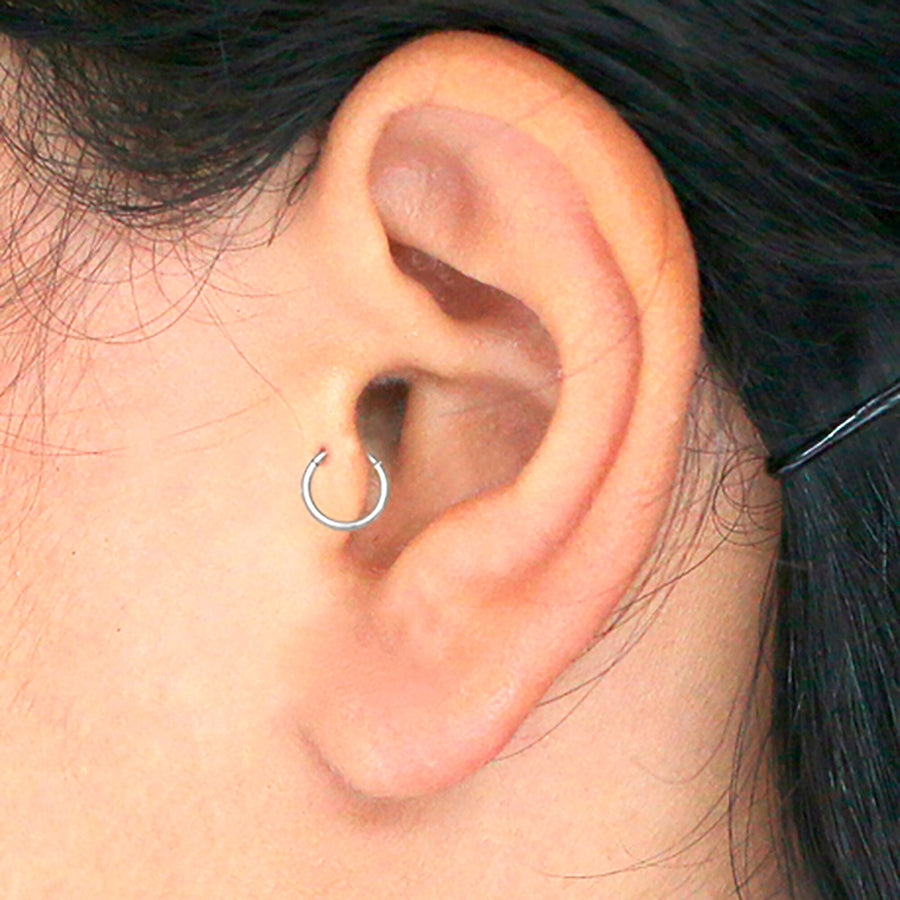 Close-up of a person's ear adorned with a Kaya Movement 316L surgical steel hoop earring in the earlobe. The black hair, partially visible, is held back by a thin black hairpin. The skin tone appears light.