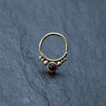 The Kaya Movement Garnet Teardrop seamless 8mm gold septum ring features a small, elegant design with a rich, dark red garnet stone at its center, encircled by delicate gold beads. The nose ring is displayed on a textured dark grey surface.