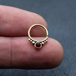 A hand holding the "Garnet Teardrop septum" ring by Kaya Movement, featuring a central red gemstone surrounded by smaller gold beads, perfectly capturing the elegance of this 8mm Gold Seamless nose ring. The background is a dark, textured surface.