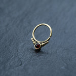 A gold nose ring, adorned with a garnet gemstone and decorative gold beads, rests on a textured dark gray surface. The circular Garnet Teardrop septum ring from Kaya Movement has an open gap at one end and features a prominent round red gem in the center flanked by small gold beads, making it a striking piece of brass body jewelry suitable for seamless nose rings, rook piercings, or daith piercings.
