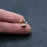 A hand holding a small, golden Garnet Teardrop septum ring by Kaya Movement, featuring a red garnet gemstone at its center. This 8mm gold septum ring, reminiscent of intricate brass body jewelry, showcases delicate gold beads accenting the top and surrounding the central gemstone. The background is a dark, textured surface.
