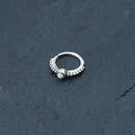 A Kaya Movement 925 silver nose ring with an iridescent opal gemstone at its center, placed on a dark, textured surface. The 8mm septum ring features a delicate band with small, evenly spaced accents leading up to the gemstone—a stunning piece of rook, tragus, or helix jewelry perfect for piercings.