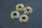 Four intricately designed Gold brest buttons piercing jewelry rings from Kaya Movement are arranged on a dark, slate surface. Each ring, resembling a delicate breast button shield, features a circular hole in the center and showcases an ornate floral motif, giving them a vintage and elegant appearance.