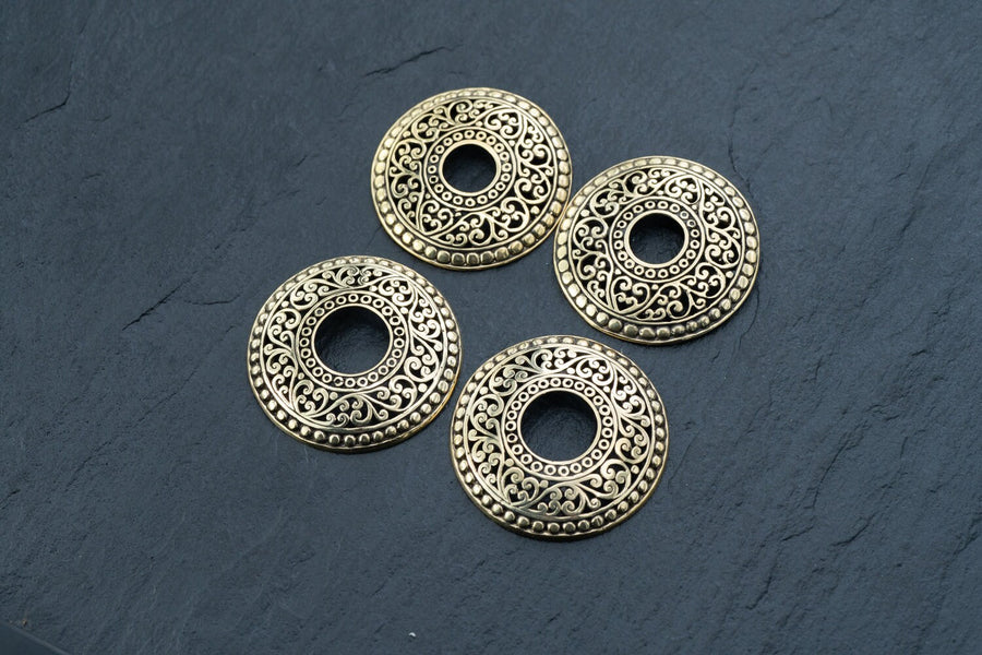 Four intricately designed Gold brest buttons piercing jewelry rings from Kaya Movement are arranged on a dark, slate surface. Each ring, resembling a delicate breast button shield, features a circular hole in the center and showcases an ornate floral motif, giving them a vintage and elegant appearance.