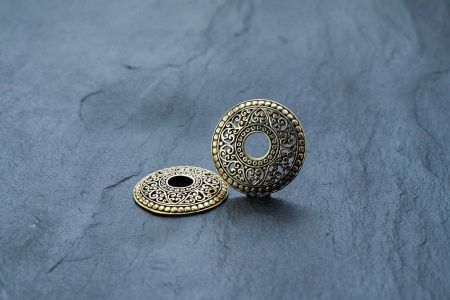 Two antiqued gold-tone, round, brass pendants with intricate filigree work lie on a slate grey surface. One pendant lies flat, showcasing its detailed circular design inspired by Kaya Movement's Gold brest buttons piercing jewelry rings and body jewelry shields, while the other stands upright, highlighting its dimension and ornate embellishments.