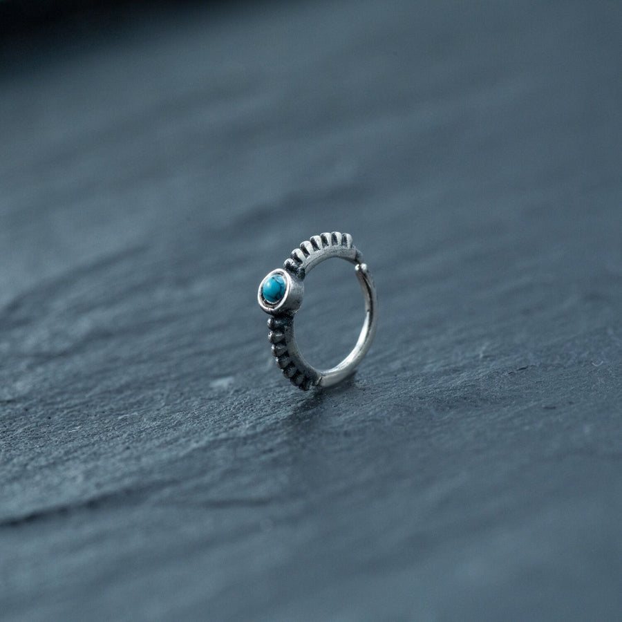 A delicate 925 silver Indian nose ring by Kaya Movement lies on a dark, textured surface. The ring features an 8mm circular turquoise stone set within a bezel, with an intricate, beaded design along the band. Perfect for septum, rook piercings, tragus piercings, or helix jewelry enthusiasts, the overall aesthetic is minimalistic yet elegant.
