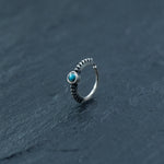 A delicate 925 silver Indian nose ring from Kaya Movement, featuring an 8mm turquoise gemstone, rests on a dark, textured surface. Reminiscent of a traditional Indian nose ring, the intricately beaded design extends halfway around the band, adding an elegant touch to this septum and helix jewelry piece.