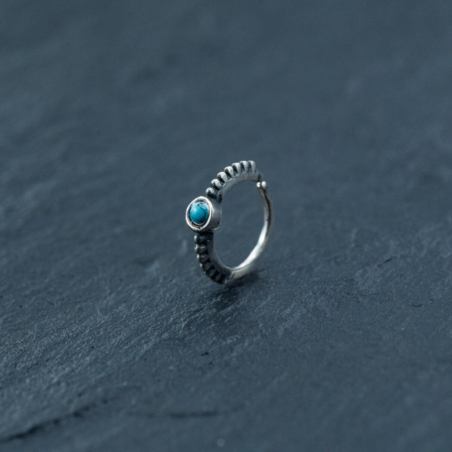 A delicate 925 silver Indian nose ring from Kaya Movement, featuring an 8mm turquoise gemstone, rests on a dark, textured surface. Reminiscent of a traditional Indian nose ring, the intricately beaded design extends halfway around the band, adding an elegant touch to this septum and helix jewelry piece.