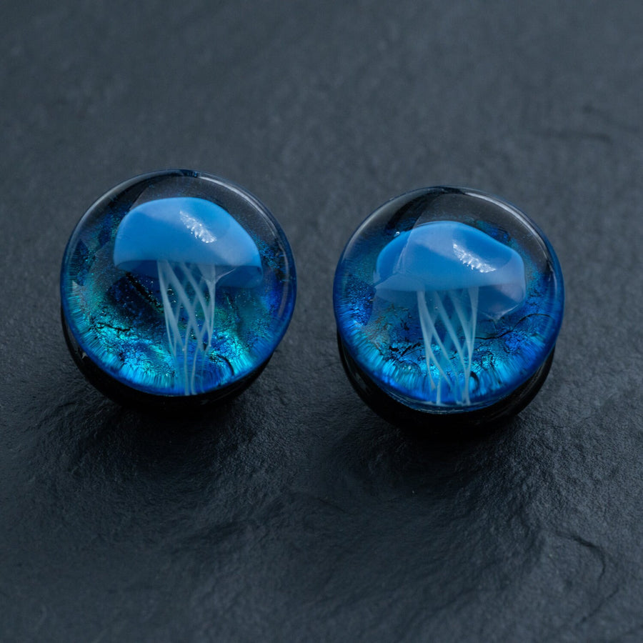 A close-up image of two rounded glass items featuring a Blue Jellyfish design encapsulated within. The jellyfish have detailed, white tentacles that stand out against the dark blue and black background of the Kaya Movement Blue Glass Jellyfish Ear Plugs - Handmade Double Flare Flesh Tunnels, 12mm-16mm. The surface on which they are placed appears to be dark and textured.