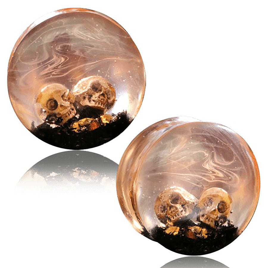 The Handmade Epoxy Double Flare Flesh Tunnels with Skull and Gothic Design by Kaya Movement feature two round plugs encapsulating small skulls in clear, swirling resin on a base of dark material adorned with gold flecks. This gauged ear stretching jewelry, available in sizes from 12mm to 16mm, exudes a haunting yet artistic appearance, making it uniquely striking.