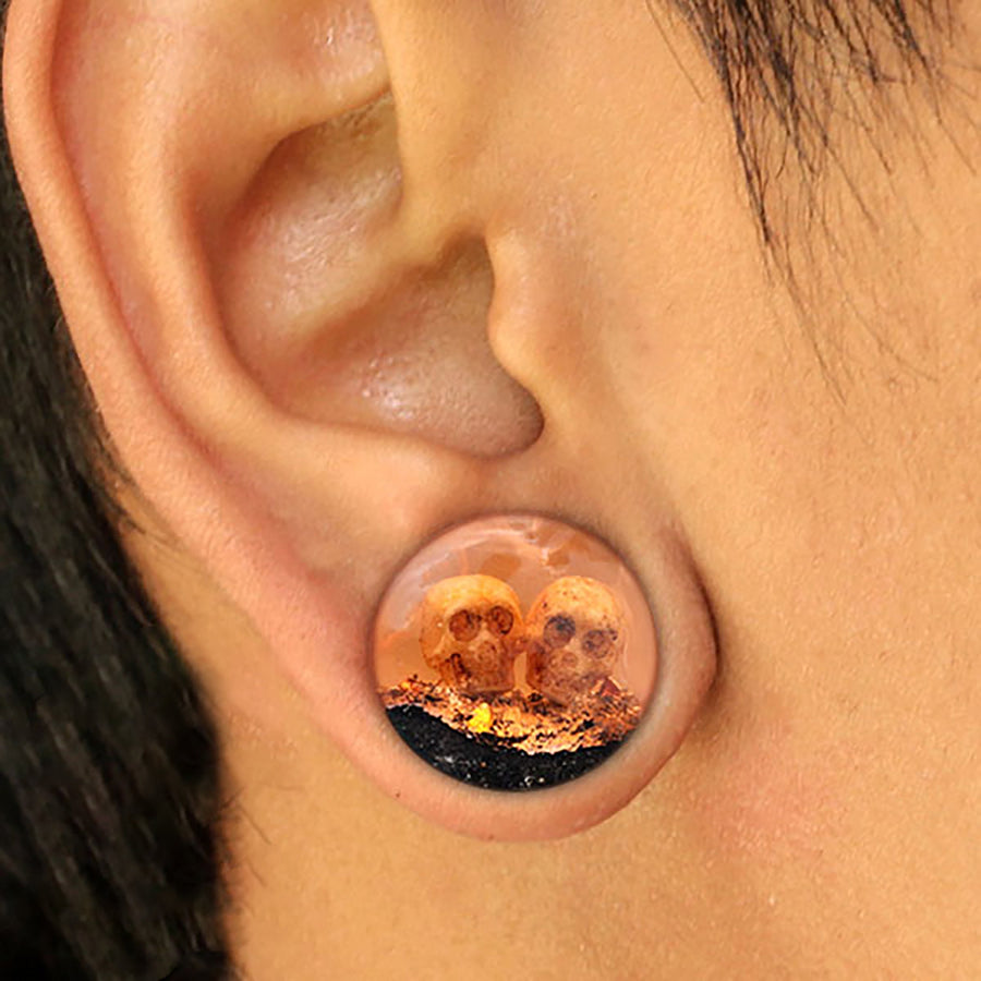 A close-up of a person's ear with a large gauge piercing, featuring the Handmade Epoxy Double Flare Flesh Tunnels by Kaya Movement. The flesh tunnel is clear, containing small skulls and a textured, gold-colored substance inside, embodying unique and intricate Gothic design. Available in 12mm-16mm gauges.