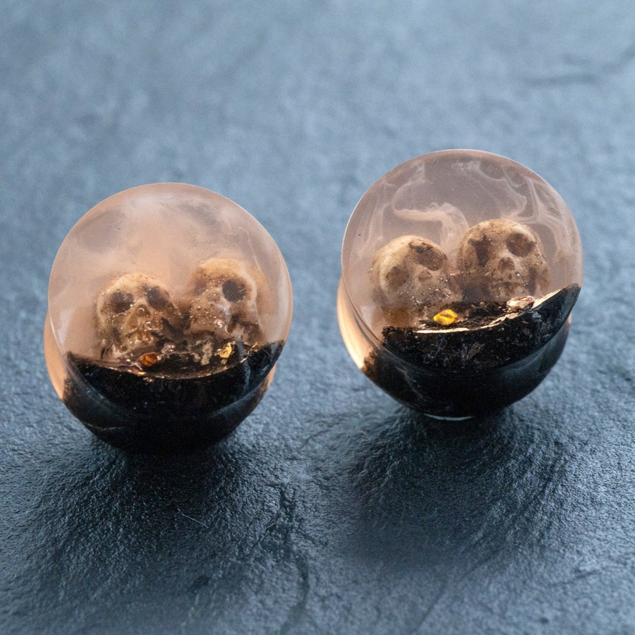 Introducing the Kaya Movement Handmade Epoxy Double Flare Flesh Tunnels with Skull and Gothic Design, available in 12mm-16mm gauges. These striking pieces of gothic jewelry feature clear resin earrings with small skulls embedded inside a gradient effect that transitions from transparent to a darker base. The skulls are partly embedded in a black layer at the bottom, making these earrings an eye-catching addition to any collection.