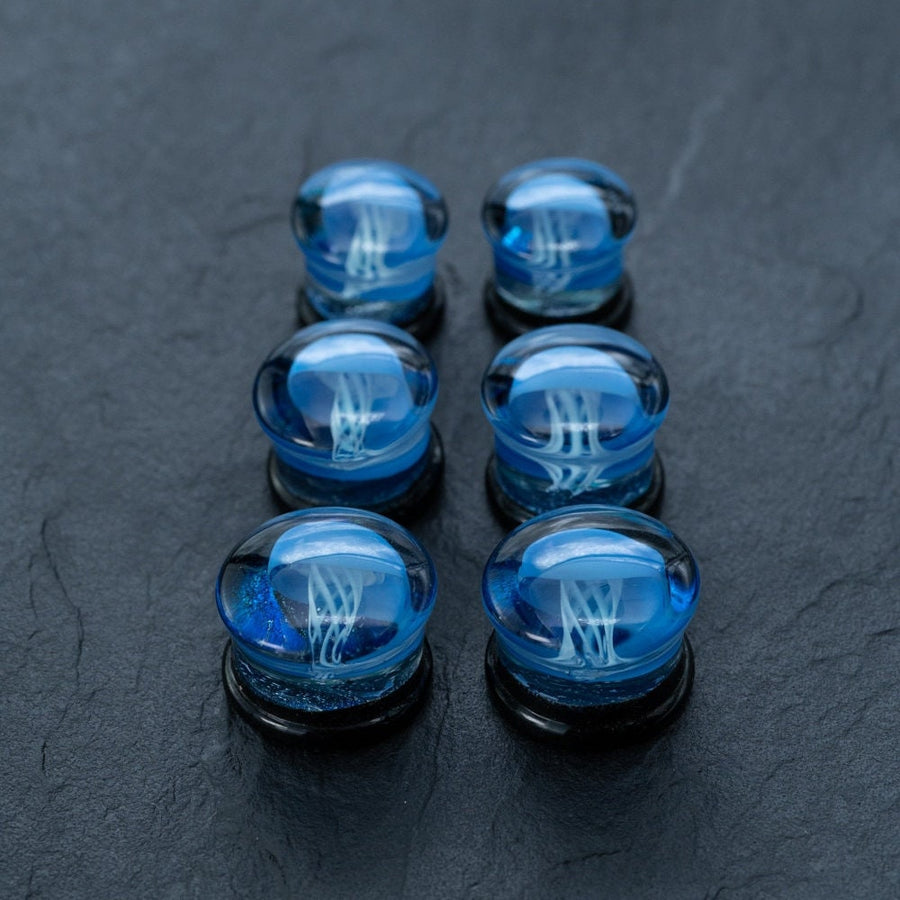 Six small, clear glass marbles featuring intricate blue jellyfish designs inside are arranged in two vertical columns on a dark, textured surface. Each marble is slightly elevated on a black base, reminiscent of the Blue Glass Jellyfish Ear Plugs by Kaya Movement. These handmade double flare flesh tunnels, available in 12mm-16mm sizes, highlight their unique ocean-themed body jewelry appeal.