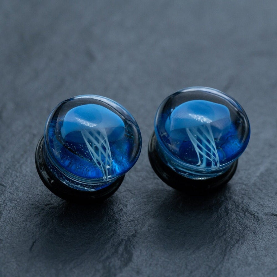 A pair of Blue Glass Jellyfish Ear Plugs by Kaya Movement, featuring round, transparent tops adorned with a vibrant blue jellyfish design inside. The handmade double flare flesh tunnels highlight a stunning jellyfish with tentacles set against a dark background, creating the illusion of it floating underwater. Available in sizes 12mm-16mm, these unique ocean-themed body jewelry pieces are perfect for those who appreciate distinctive and artistic ear plugs.