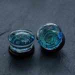 A pair of Handmade Blue Cosmic Ear Plugs from Kaya Movement, featuring round glass with mesmerizing blue and green glittery swirls that evoke galaxy or cosmic patterns. These unique celestial body jewelry pieces, perfect for stretching, are showcased against a dark, slate-like surface.