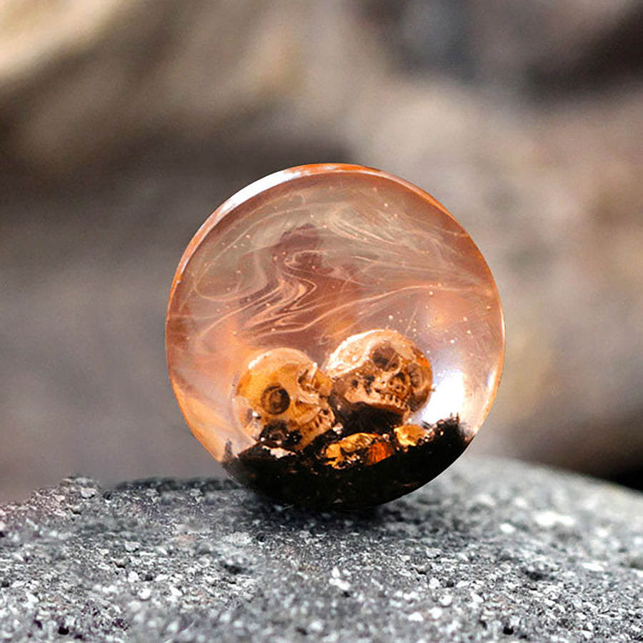 The Kaya Movement Handmade Epoxy Double Flare Flesh Tunnels, featuring small skulls embedded in dark material at the base, are crafted from a round, translucent design. Set against a blurred, rocky background, this gothic gauged ear stretching jewelry (available in 12mm-16mm) exudes an eerie and mysterious appearance.