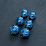 A set of six Blue Glass Jellyfish Ear Plugs by Kaya Movement, featuring miniature blue jellyfish designs encased within the round plugs, displayed on a dark, textured surface. These unique ocean-themed double flare flesh tunnels are handmade from glass and come in three progressively larger sizes ranging from 12mm to 16mm.