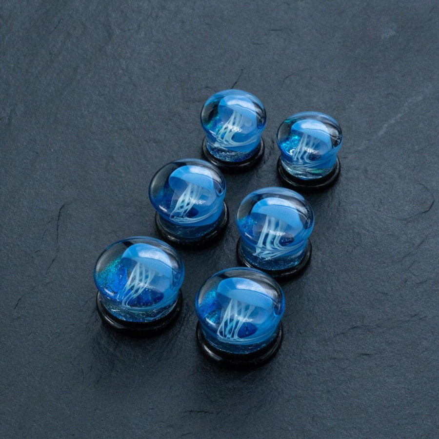 A set of six Blue Glass Jellyfish Ear Plugs by Kaya Movement, featuring miniature blue jellyfish designs encased within the round plugs, displayed on a dark, textured surface. These unique ocean-themed double flare flesh tunnels are handmade from glass and come in three progressively larger sizes ranging from 12mm to 16mm.