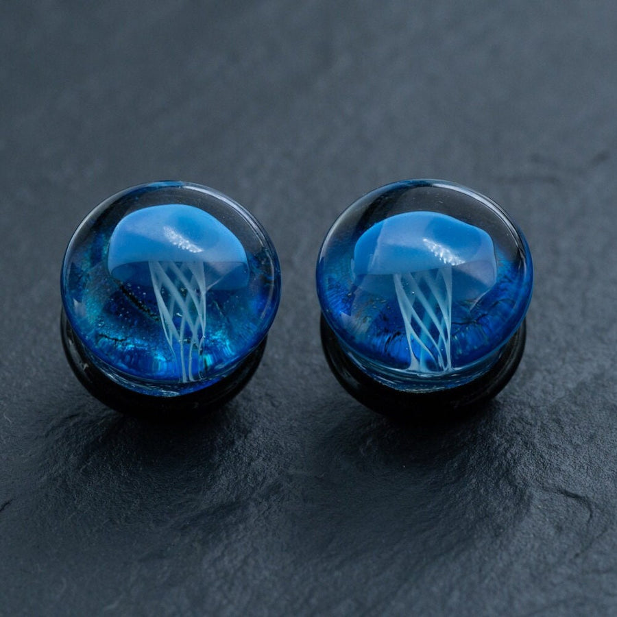 A pair of Kaya Movement handmade double flare flesh tunnels, available in sizes 12mm-16mm and featuring a blue jellyfish encapsulated in clear resin. These Blue Glass Jellyfish Ear Plugs showcase the jellyfish with its blue dome and white tentacles floating against a deep blue background, creating a unique ocean-themed aesthetic perfect for body jewelry enthusiasts.