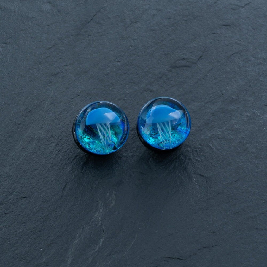 The Blue Glass Jellyfish Ear Plugs by Kaya Movement feature an intricate Blue Jellyfish design encased inside small, round plugs. The jellyfish appear to float in a deep, translucent blue resin, creating a captivating 3D effect against a dark grey textured background. These handmade double flare flesh tunnels are available in sizes ranging from 12mm to 16mm and make stunning ocean-themed body jewelry that captivates and enchants.