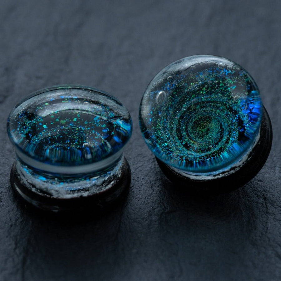 A pair of Kaya Movement's Handmade Blue Cosmic Ear Plugs, featuring swirling blue and green patterns with glitter suspended inside, are displayed on a dark, textured surface. These round, transparent Spiral Gauges (Double Flare Tunnels) in sizes 12mm-16mm resemble miniature galaxies encased in glass, making for unique celestial body jewelry for stretching.