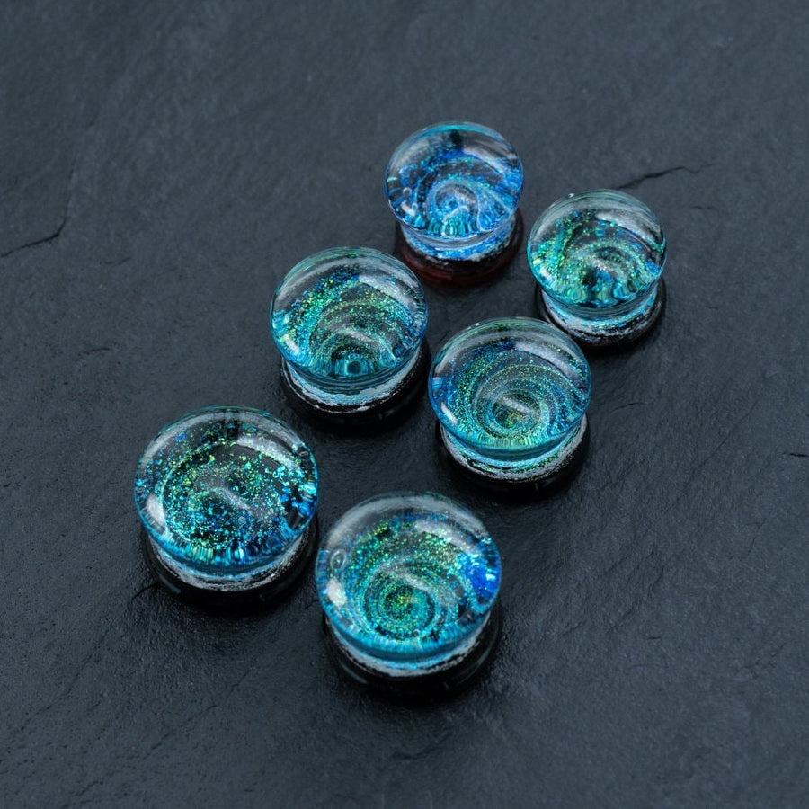 A set of six Handmade Blue Cosmic Ear Plugs by Kaya Movement, each featuring intricate swirling patterns of blue, green, and white that resemble glass marbles. Resting on a dark gray, textured surface, these unique celestial body jewelry pieces showcase mesmerizing galaxy-like designs. Available in 12mm-16mm sizes with spiral gauges and double flare tunnels for stretching.
