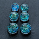 A close-up of six iridescent Handmade Blue Cosmic Ear Plugs from Kaya Movement arranged in two columns on a dark surface. The plugs, ranging from 12mm to 16mm, boast swirling patterns of blue, green, and gold, resembling miniature galaxies or ocean waves within them.