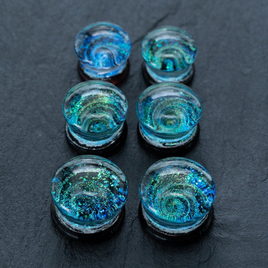 A close-up of six iridescent Handmade Blue Cosmic Ear Plugs from Kaya Movement arranged in two columns on a dark surface. The plugs, ranging from 12mm to 16mm, boast swirling patterns of blue, green, and gold, resembling miniature galaxies or ocean waves within them.