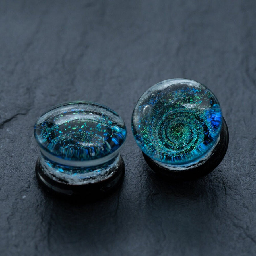 Close-up image of two Handmade Blue Cosmic Ear Plugs from Kaya Movement, featuring intricate blue and green galaxy-like patterns encased within. These unique celestial body jewelry pieces are set against a dark, slate-like background, highlighting their shimmering details and colors. The Spiral Gauges, Double Flare Tunnels (available in 12mm-16mm) are truly mesmerizing for those looking to stretch their piercings in style.