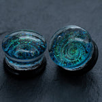 The Kaya Movement’s Handmade Blue Cosmic Ear Plugs are a striking piece of body jewelry, featuring black, handmade ear plugs encased in glass domes. Each plug sports a mesmerizing blue and green swirling design that resembles a galaxy, standing out vividly against the dark, textured backdrop. These unique celestial double flare tunnels come in sizes ranging from 12mm to 16mm and are perfect for stretching.