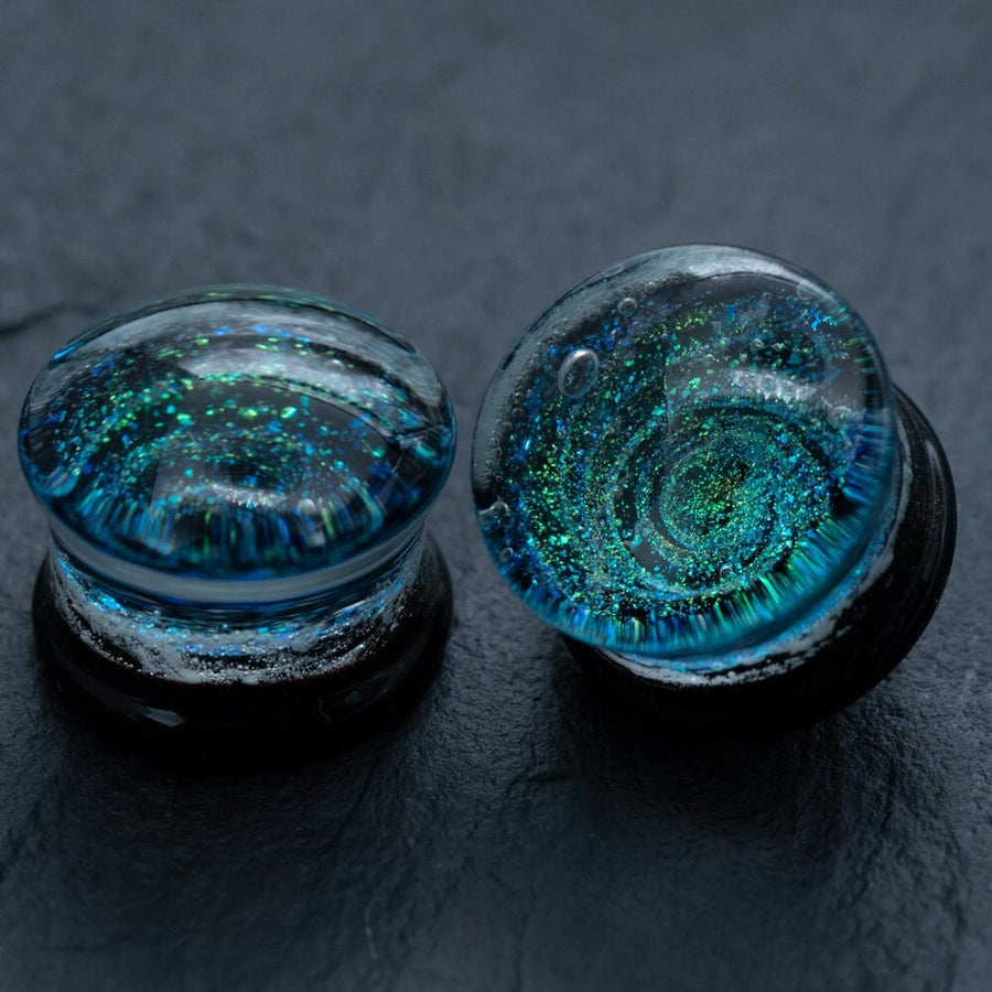 The Kaya Movement’s Handmade Blue Cosmic Ear Plugs are a striking piece of body jewelry, featuring black, handmade ear plugs encased in glass domes. Each plug sports a mesmerizing blue and green swirling design that resembles a galaxy, standing out vividly against the dark, textured backdrop. These unique celestial double flare tunnels come in sizes ranging from 12mm to 16mm and are perfect for stretching.