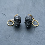 Product Description: Introducing the Handmade Black Skull Dangle Earrings from Kaya Movement, a stunning piece of gothic tribal jewelry designed for a bold statement style. These earrings feature intricately detailed black skulls accented with gold-tone rings attached at the top, all set against a dark, textured surface. The contrast between the slightly shiny black skulls and the bright gold rings adds an eye-catching element to this unique and stylish accessory.