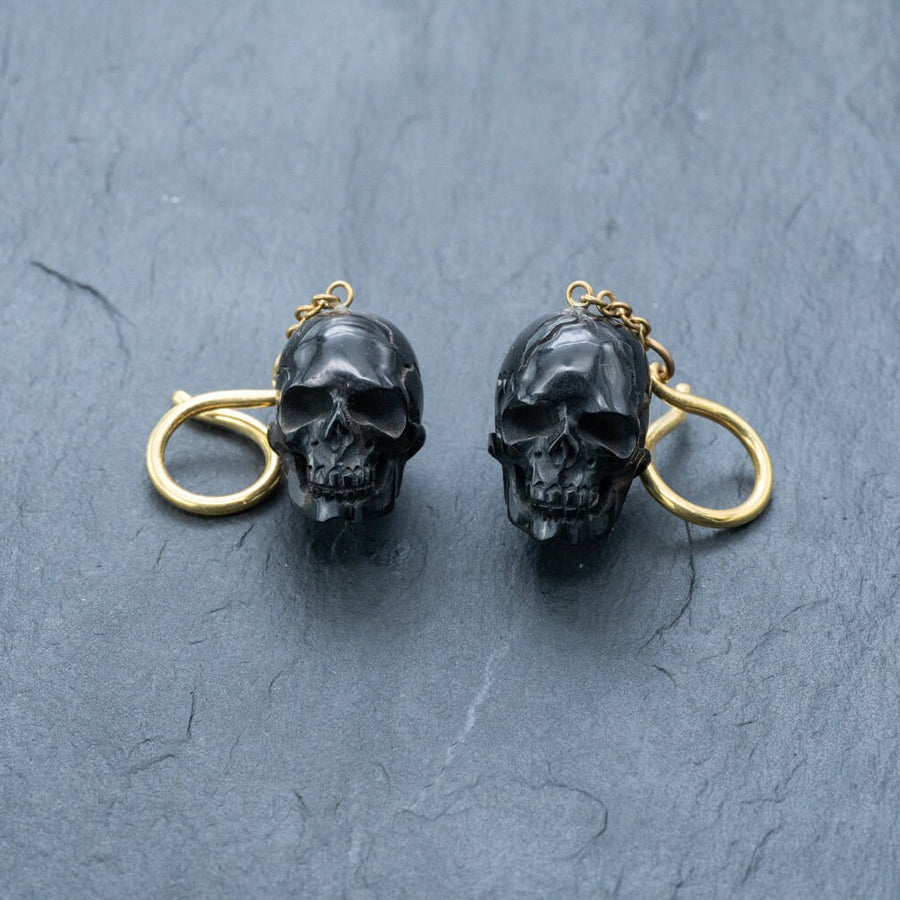 Product Description: Introducing the Handmade Black Skull Dangle Earrings from Kaya Movement, a stunning piece of gothic tribal jewelry designed for a bold statement style. These earrings feature intricately detailed black skulls accented with gold-tone rings attached at the top, all set against a dark, textured surface. The contrast between the slightly shiny black skulls and the bright gold rings adds an eye-catching element to this unique and stylish accessory.