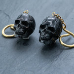 The Handmade Black Skull Dangle Earrings by Kaya Movement feature dark metallic skull pendants with intricate detailing. The black skulls are attached to golden-colored loops, creating a striking contrast. Placed on a dark, textured surface, these earrings are ideal for any gothic tribal jewelry collection and perfect for making a bold statement.