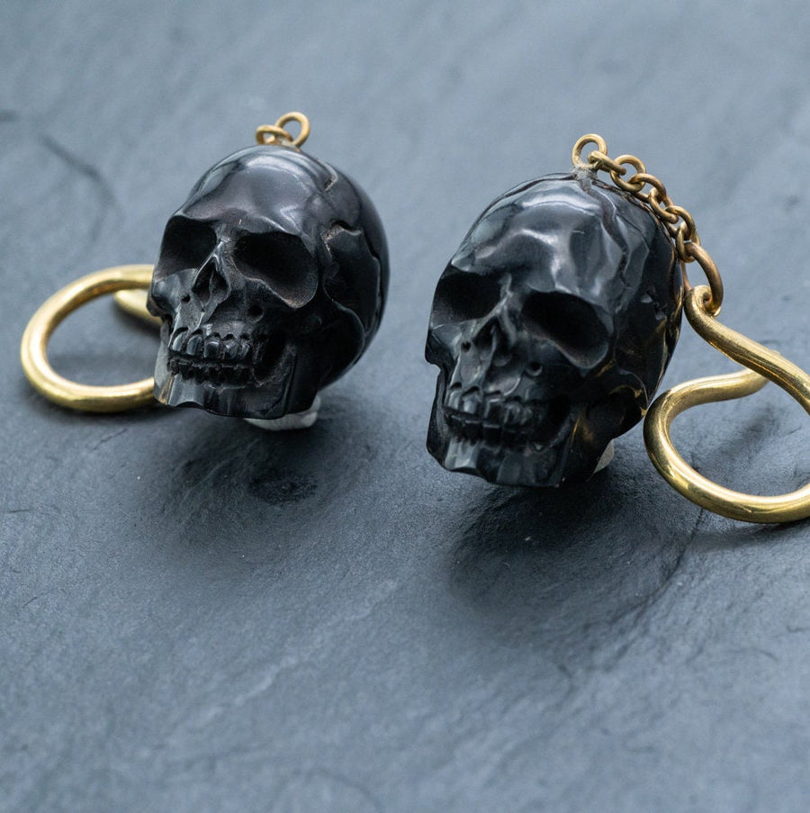 The Handmade Black Skull Dangle Earrings by Kaya Movement feature dark metallic skull pendants with intricate detailing. The black skulls are attached to golden-colored loops, creating a striking contrast. Placed on a dark, textured surface, these earrings are ideal for any gothic tribal jewelry collection and perfect for making a bold statement.