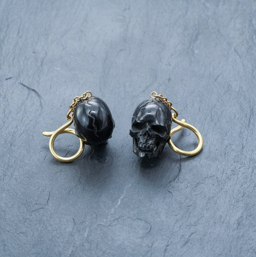 The Handmade Black Skull Dangle Earrings by Kaya Movement feature skull-shaped pendants that hang from gold hooks, set against a textured gray surface. The intricate details of this gothic tribal jewelry are highlighted by the contrasting dark hue of the black skulls against the gold.