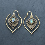 A pair of intricately designed unisex handmade brass leaf ear weights from Kaya Movement rested on a dark surface. These boho hippie tribal earrings are teardrop-shaped with detailed patterns, featuring a small, iridescent abalone shell set in the center. The outer part elegantly forms an eye-catching loop, perfect for edgy urban fashion and gauged dangle gold earring styles.