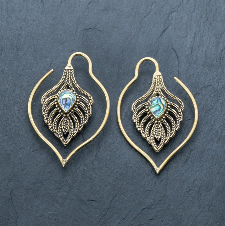 A pair of intricately designed unisex handmade brass leaf ear weights from Kaya Movement rested on a dark surface. These boho hippie tribal earrings are teardrop-shaped with detailed patterns, featuring a small, iridescent abalone shell set in the center. The outer part elegantly forms an eye-catching loop, perfect for edgy urban fashion and gauged dangle gold earring styles.