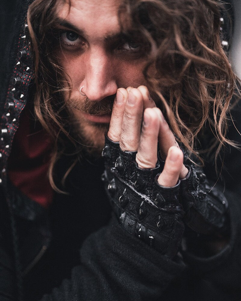 A person with long, wavy hair and a nose ring gazes intently at the camera. They are wearing Kaya Movement's Custom Dragon Scale Leather Gloves, which are fingerless motorcycle and driving gloves perfect for post-apocalyptic goth stagewear. Along with a hooded jacket adorned with metallic studs, their hands partially cover their face, adding a mysterious and intense expression that hints at gothic fashion influences.