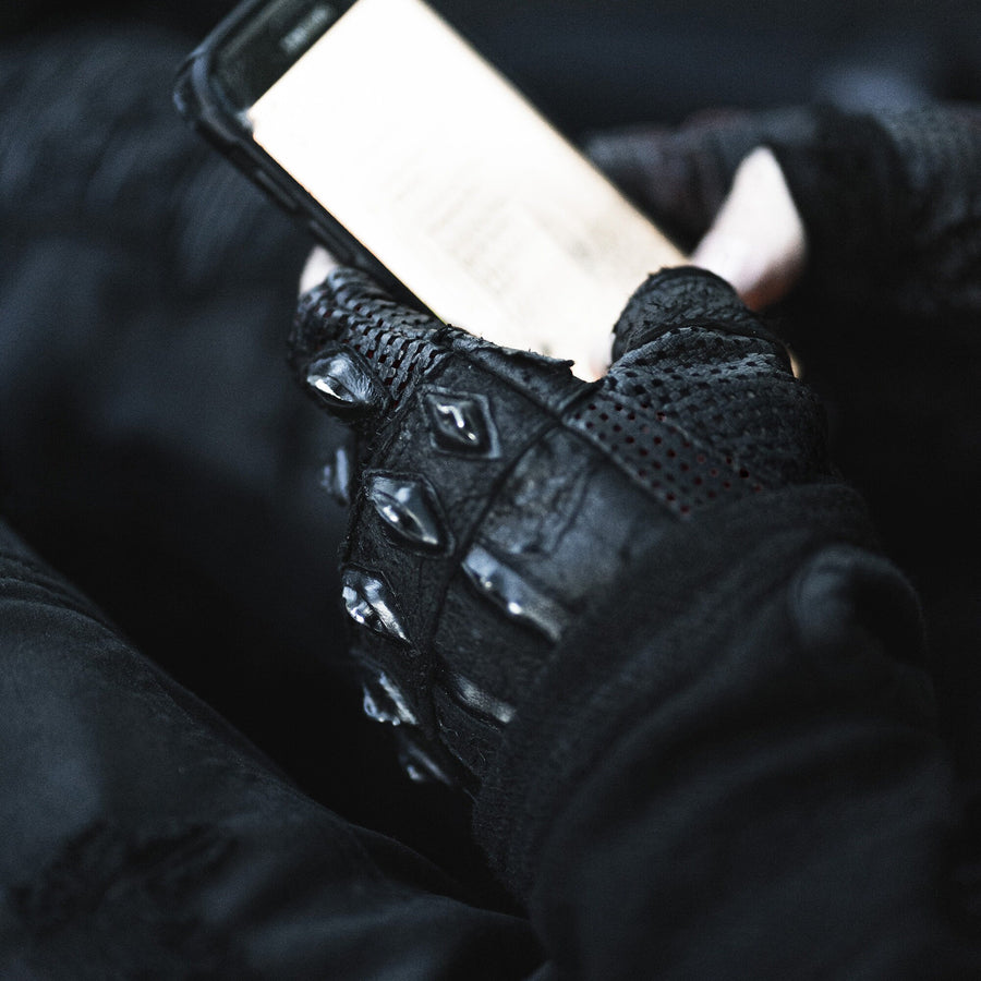A person wearing Kaya Movement's Custom Dragon Scale Leather Gloves, Fingerless Motorcycle & Driving Gloves, stylish biking and gothic stagewear, holds a smartphone. The screen of the phone is lit, showing some text, suggesting the person is reading or typing. The gloves have a textured, armored design with raised elements for protection, appealing to both motorcycle enthusiasts and fans of post-apocalyptic goth fashion.