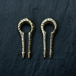 A pair of intricately designed keyhole and hanger-style gold ear weights from the Kaya Movement collection is showcased against a dark textured background. Ideal for the stretched ear game, these minimalist earrings feature engraved patterns and boast an antique appearance.